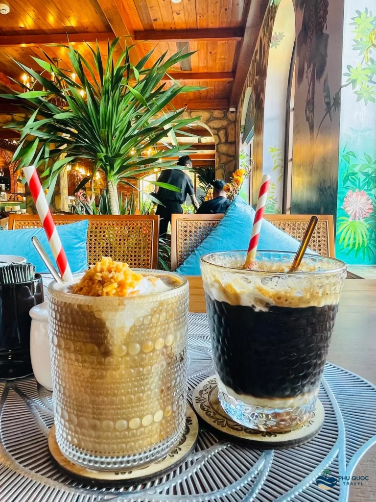 A creamy coconut coffee in a tall glass, with layers of coffee and coconut milk, sits on a rustic wooden table at Venice Cafe. The Phu Quoc sunset paints the sky orange and pink in the background.
